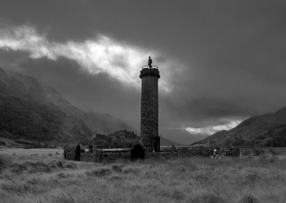 A black and white image of a cylindrical monument with a figure of a man on top. It's standing in a stone-walled circular enclosure and lies at the foot of a glen. It's a stormy day with a threatening sky.