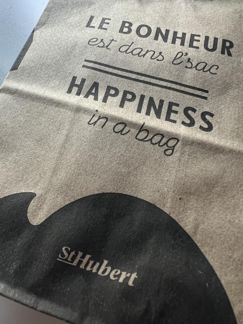 An angled view of a brown paper bag with black wording: “LE BONHEUR est dans l’sac  || HAPPINESS in a bag” with a black design and a company logo “St Hubert”. [Quebec, Canada (June 2026)]