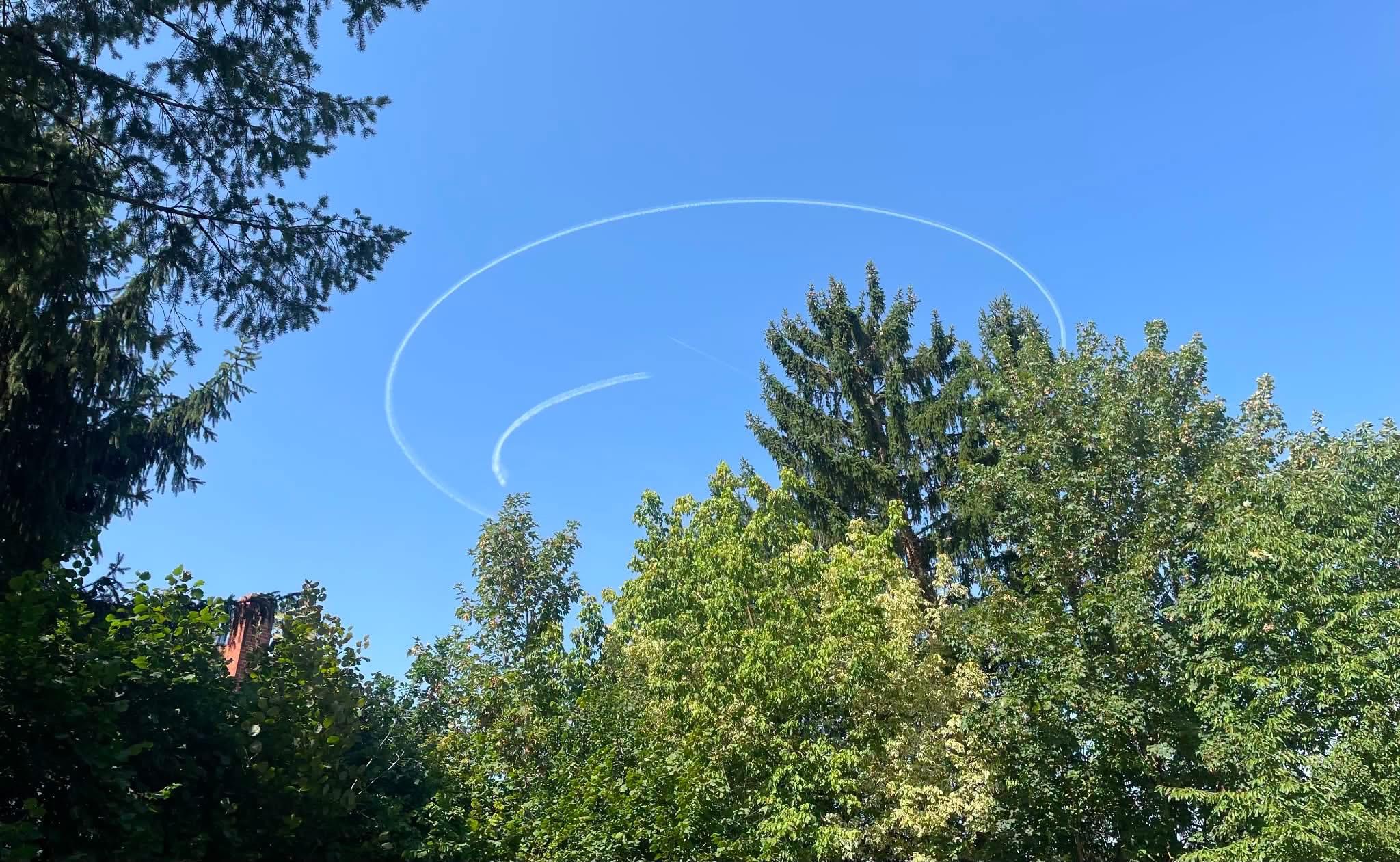 A clear blue sky filled with white contrails forming circular patterns above tall green trees.