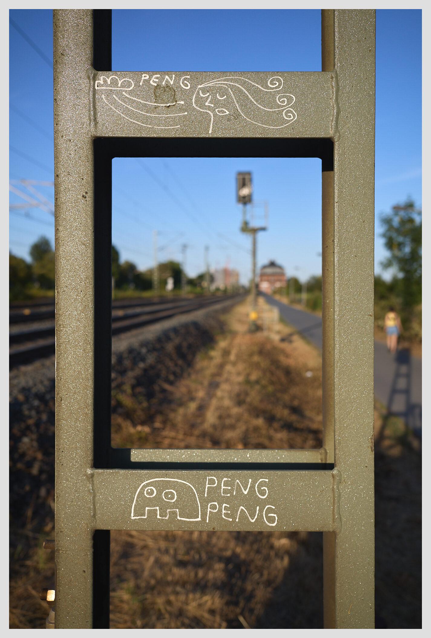 Vertical closeup of a signal tower next to a railway track. The greenish metal beams have been embellished with two small figures by local street artist peng. Through the opening in the structure the train tracks are visible on the left. On the right, a paved walkway runs parallel to the tracks. A person can be seen on the walkway. The scene is lit by warm late afternoon light.