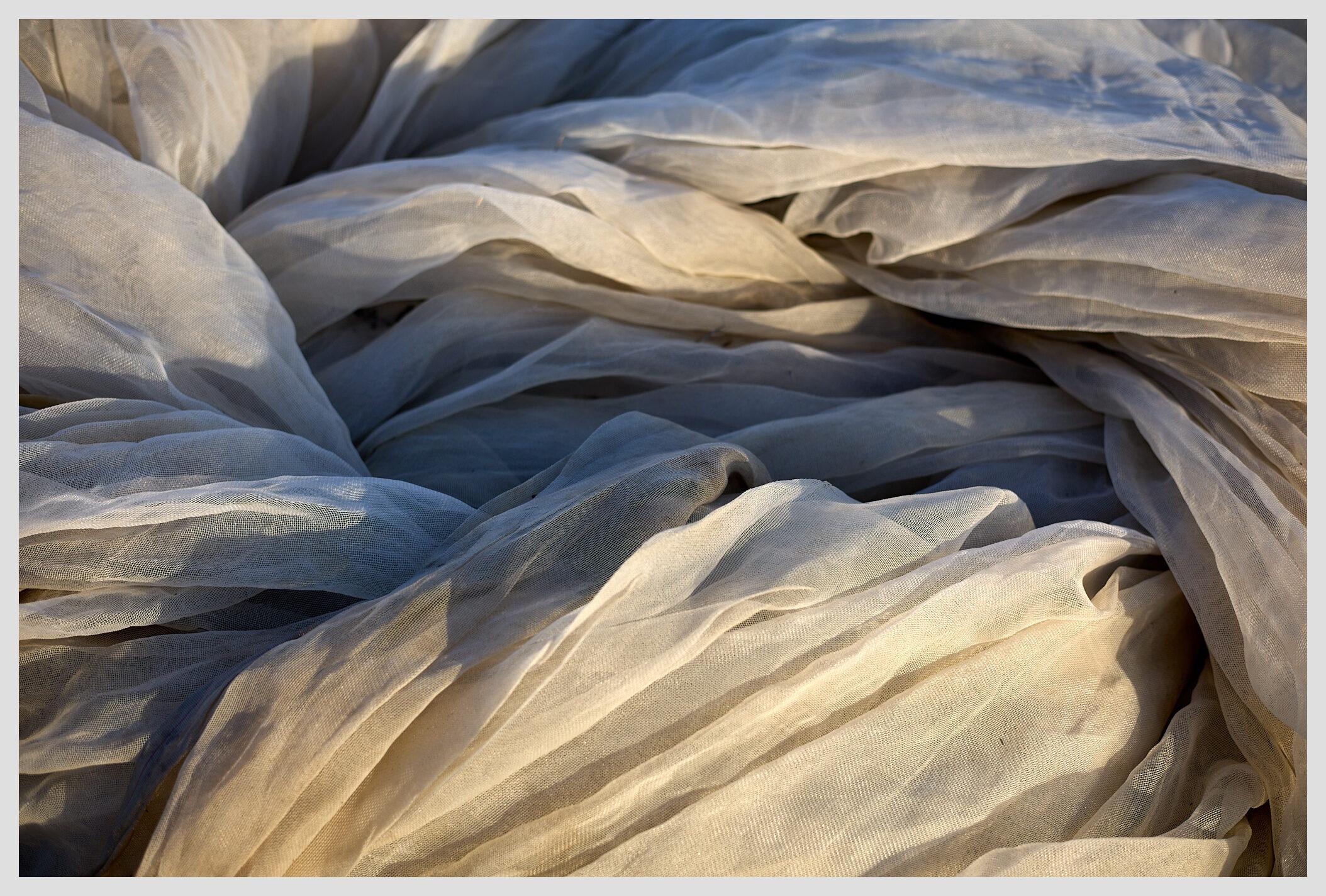 Horizontal top down closeup of a pile of white protective nets used on a field to shield the vegetables. The nets have been gathered next to a field and fill the entire frame. Warm evening light illuminates the scene from there bottom of the frame.
