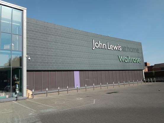 Store front. Grey metallic cladding with text "John Lewis Waitrose" with faded text reading "at home" following "John Lewis". Blue sky above. Grey/Brown tarmac in the foreground.