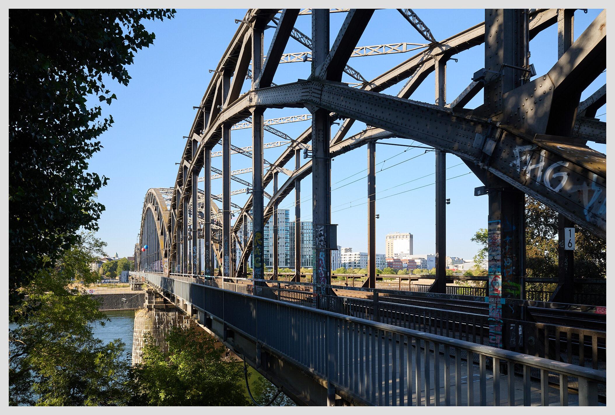 Horizontal image of the Deutschherrnbrücke, a steel train bridge in Frankfurt crossing the river Main, with a narrow walkway for pedestrians on the left. Taken from the southern bank with the camera pointed to the East end.