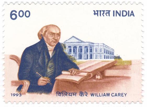 A commemorative postage stamp with an image of William Carey seated at a able with a book open in front of him. In the background serampore College building.