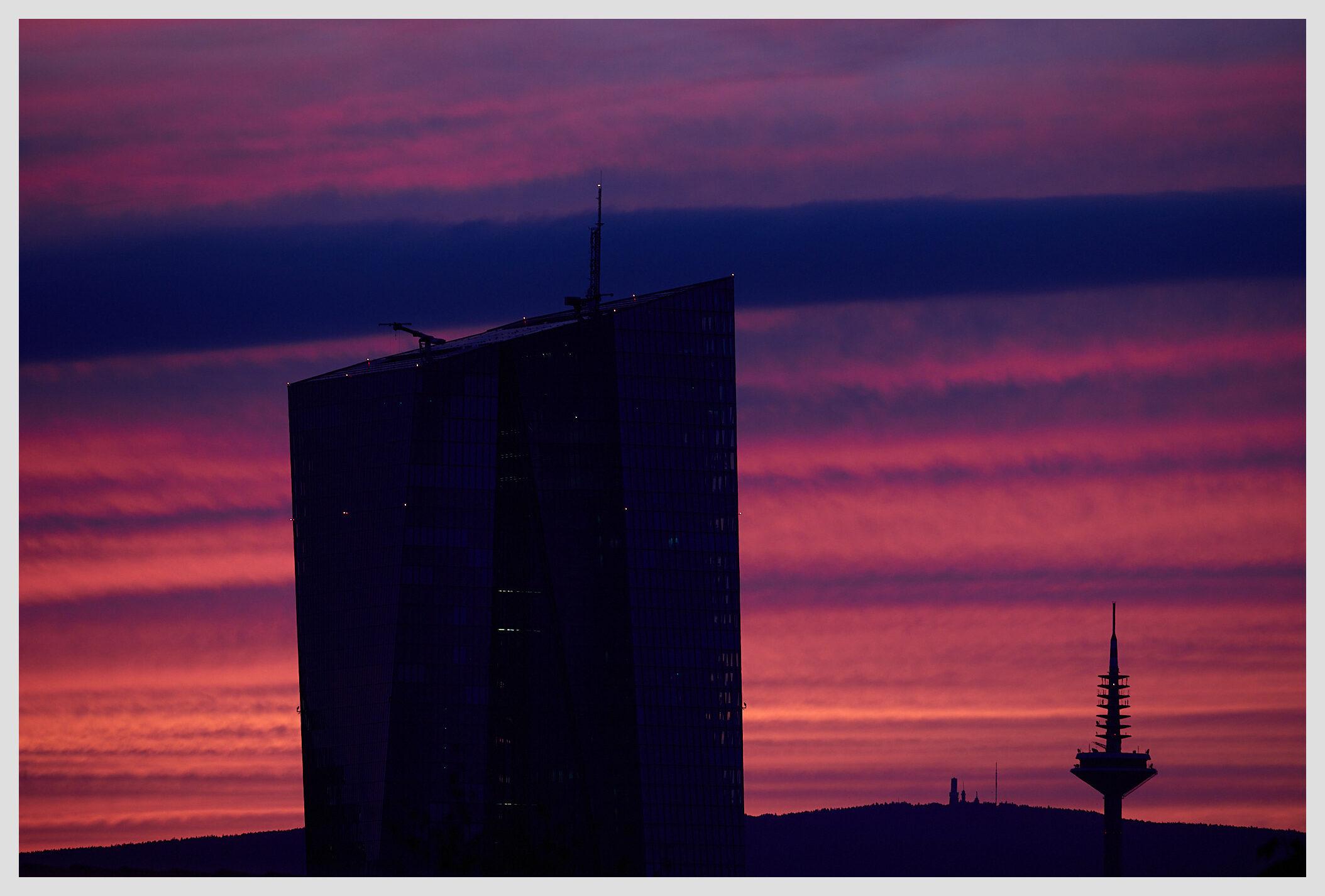 Horizontal image of a highrise in a city against a dramatic red-violet striped sunset sky. The structures are visible as silhouettes: A highrise in the foreground / center-left of the frame. A comms tower in the middleground / right of the frame, and another comms tower on a far away mountain range in the background / center of the frame.