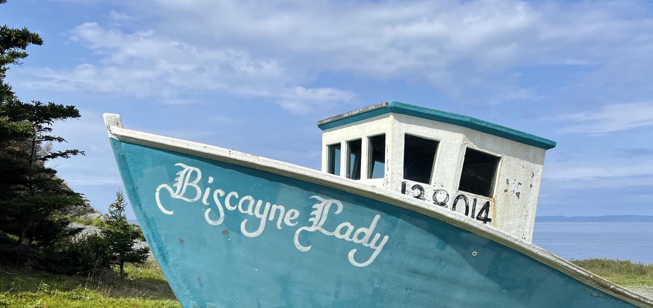 A teal fishing boat with a white cabin sits on a grassy coast with a blue sky in the background. The front of the boat has white lettering “Biscayne Lady” in a fancy script, and the cabin has an ID number painted in it in blue. An evergreen tree peeks into the left edge of the photo, while the right shows a blue bay that is very similar in colour to the partially clouded blue sky. [Bauline (NL, Canada) August 2026]