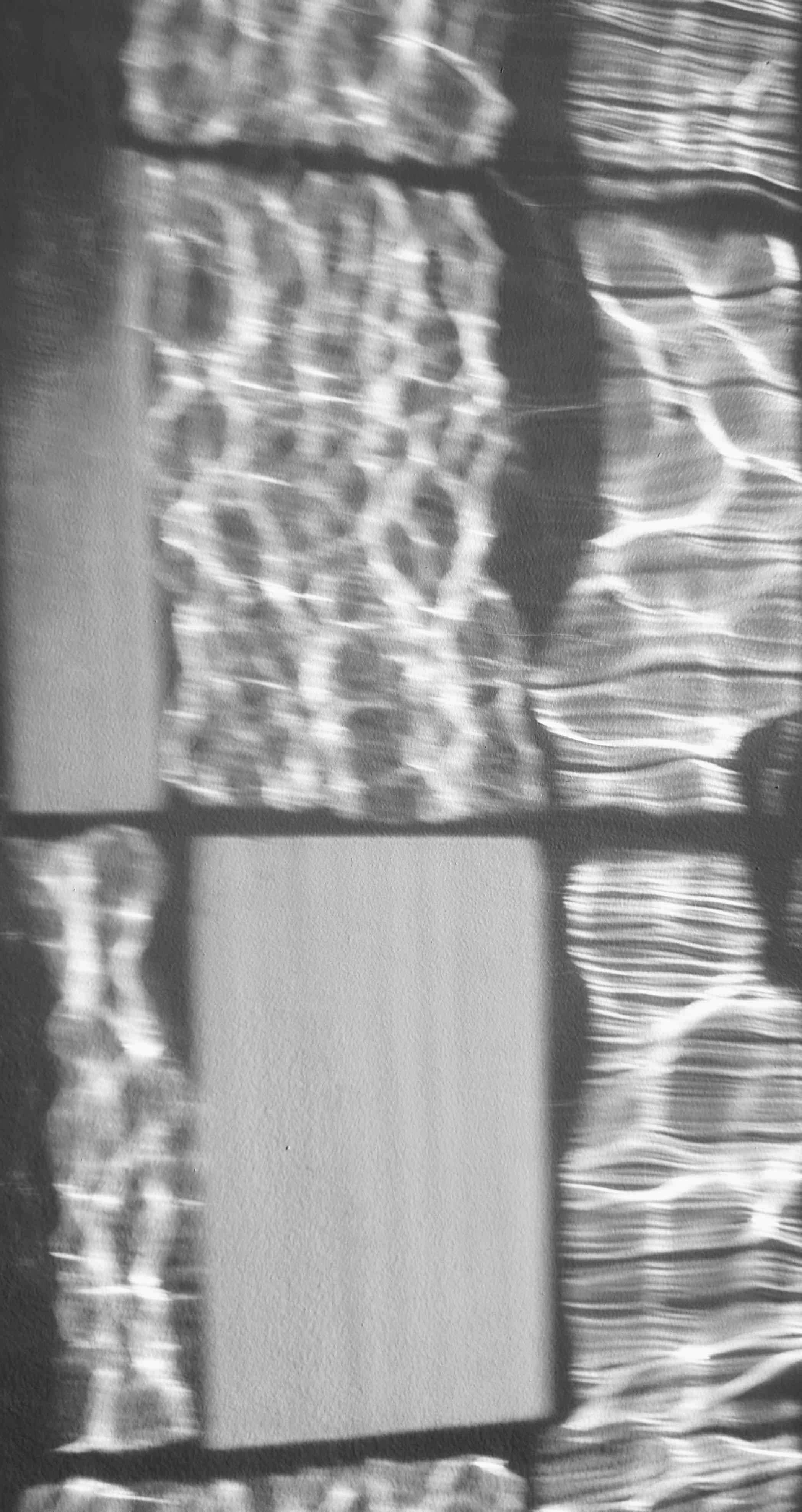 This is a black and white digital photo. It shows an abstract effect produced by sunlight shining at a low angle through a window, and projecting refraction patterns onto the opposite wall. The window is made of several rectangular panes of glass. Each pane is simply regular glass, meant for looking outside and letting light in. But in the projection effect, each pane produces its own pattern as if in a grid. The effect in each pane seems wavelike and could be mistaken for interference patterns. However the patterns just reveal and amplify imperfections in the flatness of the panes (the window could be as much as one hundred year old). The overall image seems slightly out of focus, but that is actually what it looked like on the wall where the photograph was taken.