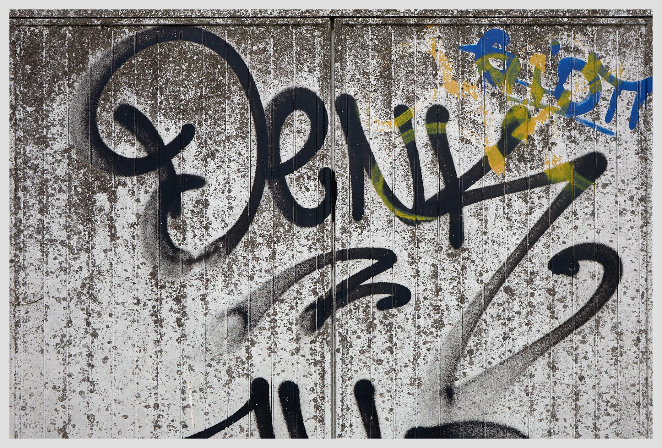 Horizontal closeup of a large 'Denk' graffiti tag on a gray switch box. The image could be mistaken for black and white if not for a smaller yellow and blue tag in the top right corner.