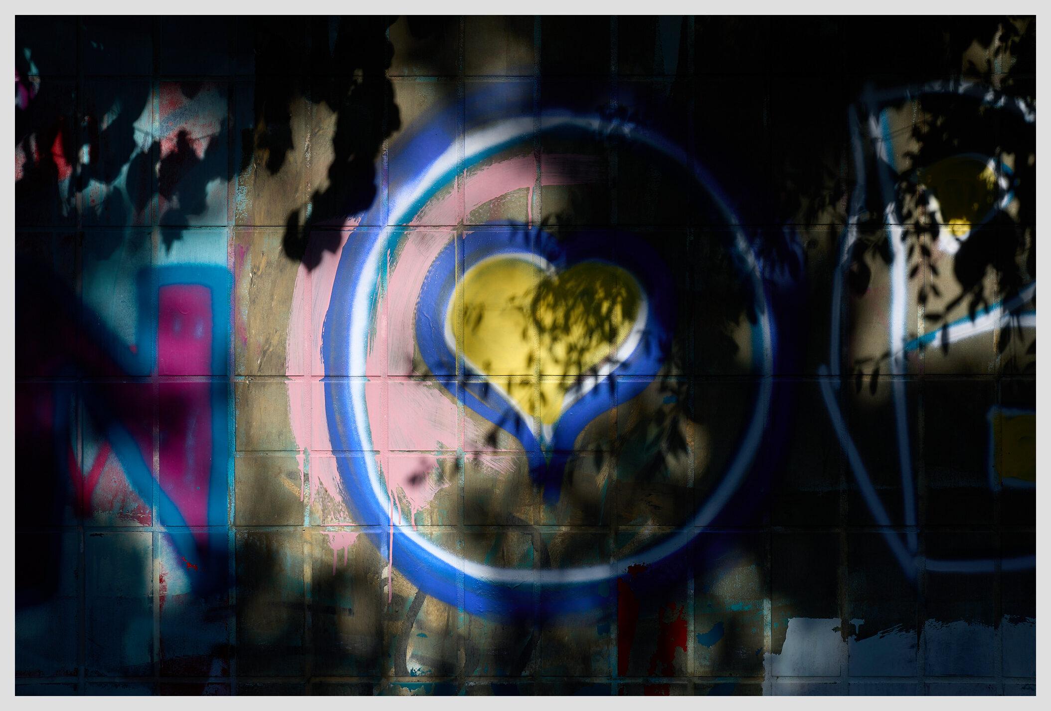 Horizontal image of a tiled wall covered in graffiti at the entrance of an underpass. A bright yellow heart is at the center of the frame, illuminated by sunlight. Shadows of foliage frame the image.