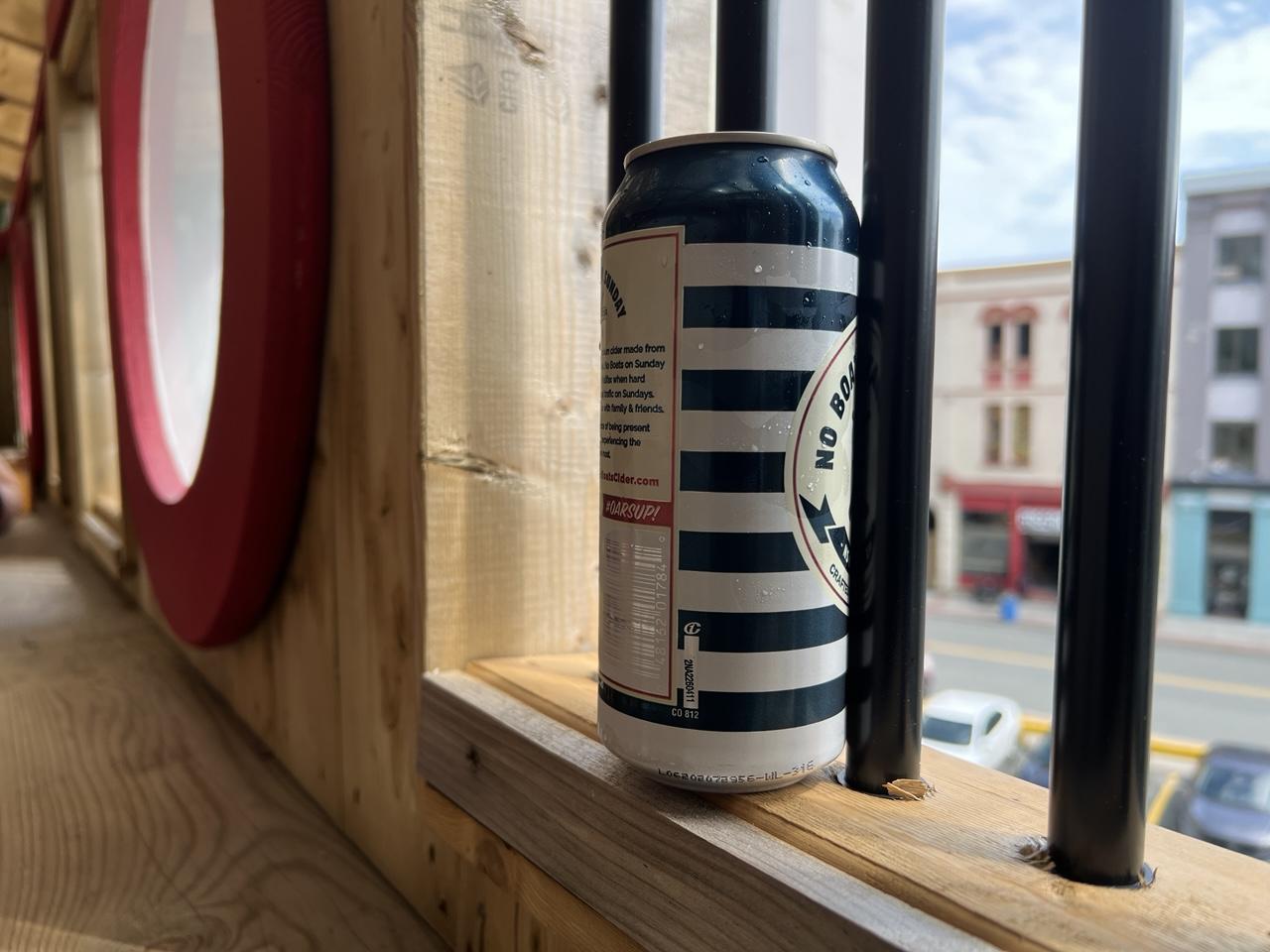 A blue and white striped beverage can sits on a black barred wooden windowsill overlooking a city street with coloured windowed buildings in the blurred background. The photo angle shows other red-framed porthole style windows blurred into the background. [The Ship (St. John’s, CA) August 2026]