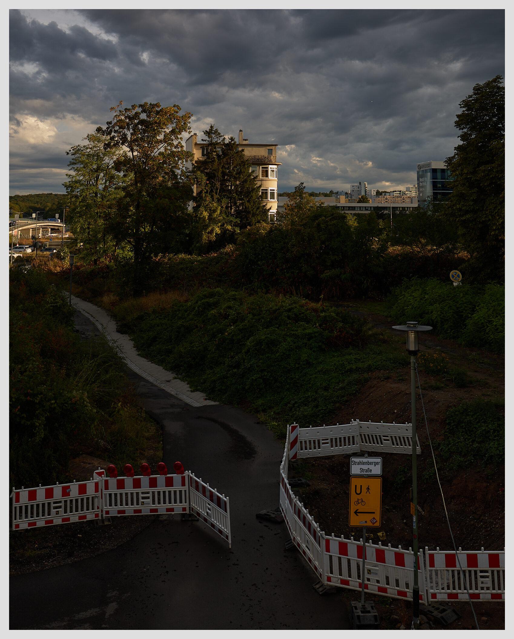Wide vertical image taken in the city from an elevated viewpoint. A lone building stands in an urban landscape, illuminated by warm sunlight on its right side. The sky is overcast with dark clouds after storm just passed.