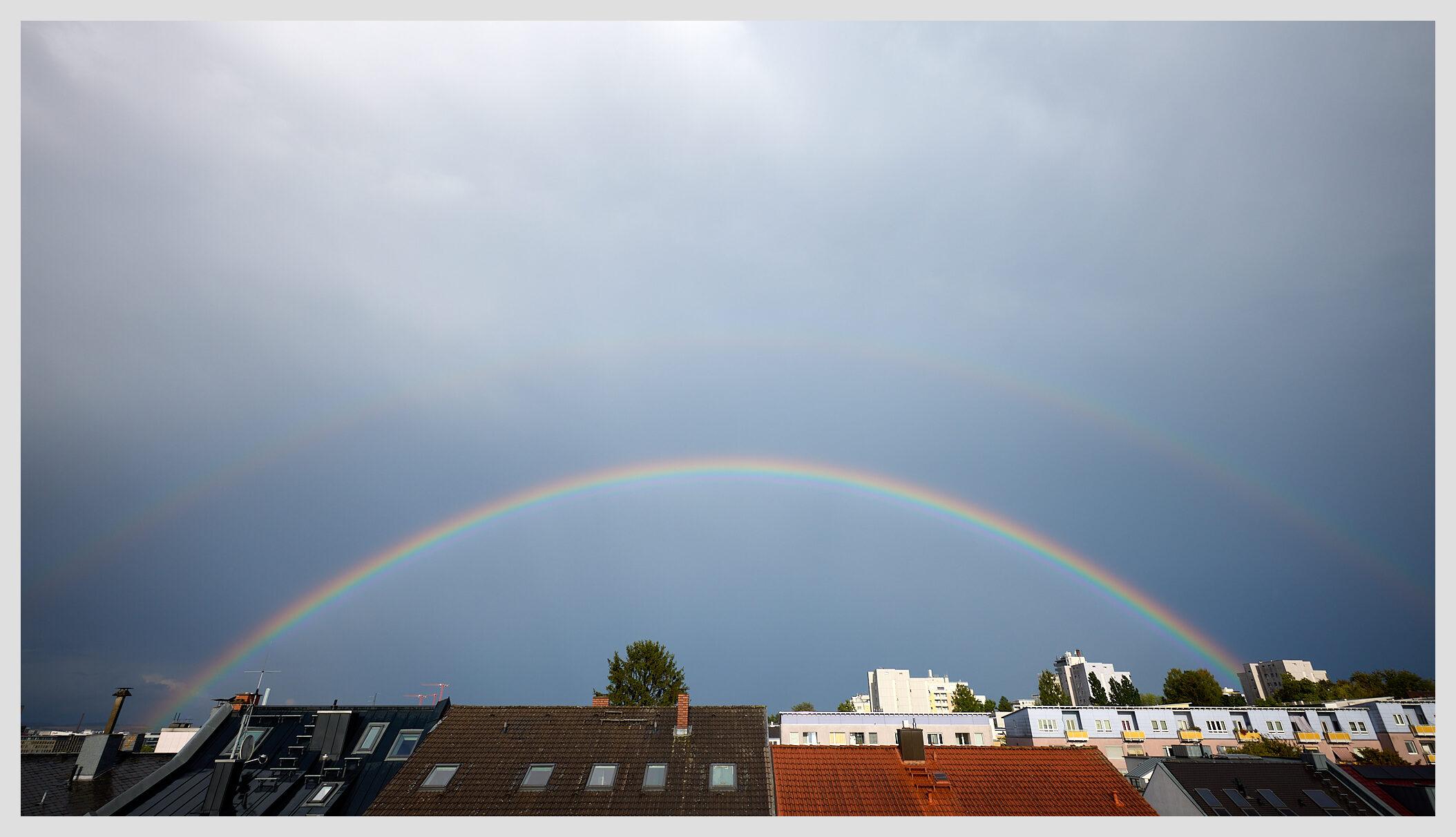 Wide panoramic image of a double rainbow over the roofs of a city. The sky is grayish-blue.