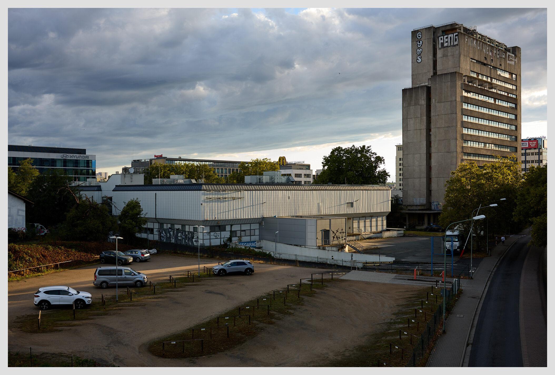 Wife horizontal image of an almost empty parking lot in a mixed office / industrial zone. On the right side of the frame, a large gray building towers over the scene. A large peng tag has been painted near the top. The sky is dark with clouds after a storm has passed through, the sunlight coming from the right gives the scene some dramatic contrast.