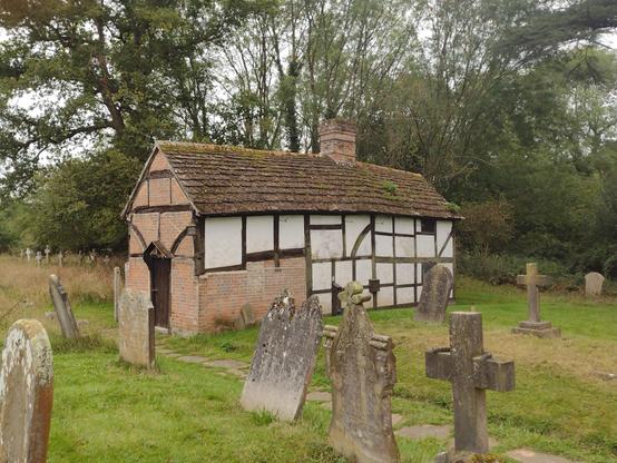 A timber-framed, mainly white and black, render and wood, single-story building with a stone roof. Set in a churchyard with trees in the background.