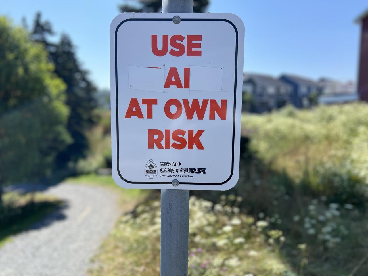 A vandalized city hiking trail sign reads « USE **AI* AT OWN RISK ».  The white rectangular sign has red letters, with a small « Grand Concourse » logo near the bottom. It’s mounted on a grey metal pole and shows a blurred grey stone path that curves around leafy green trees and a grassy field backing low apartment buildings in front of a cloudless blue sky. [St. John’s (NL, Canada) August 2026]