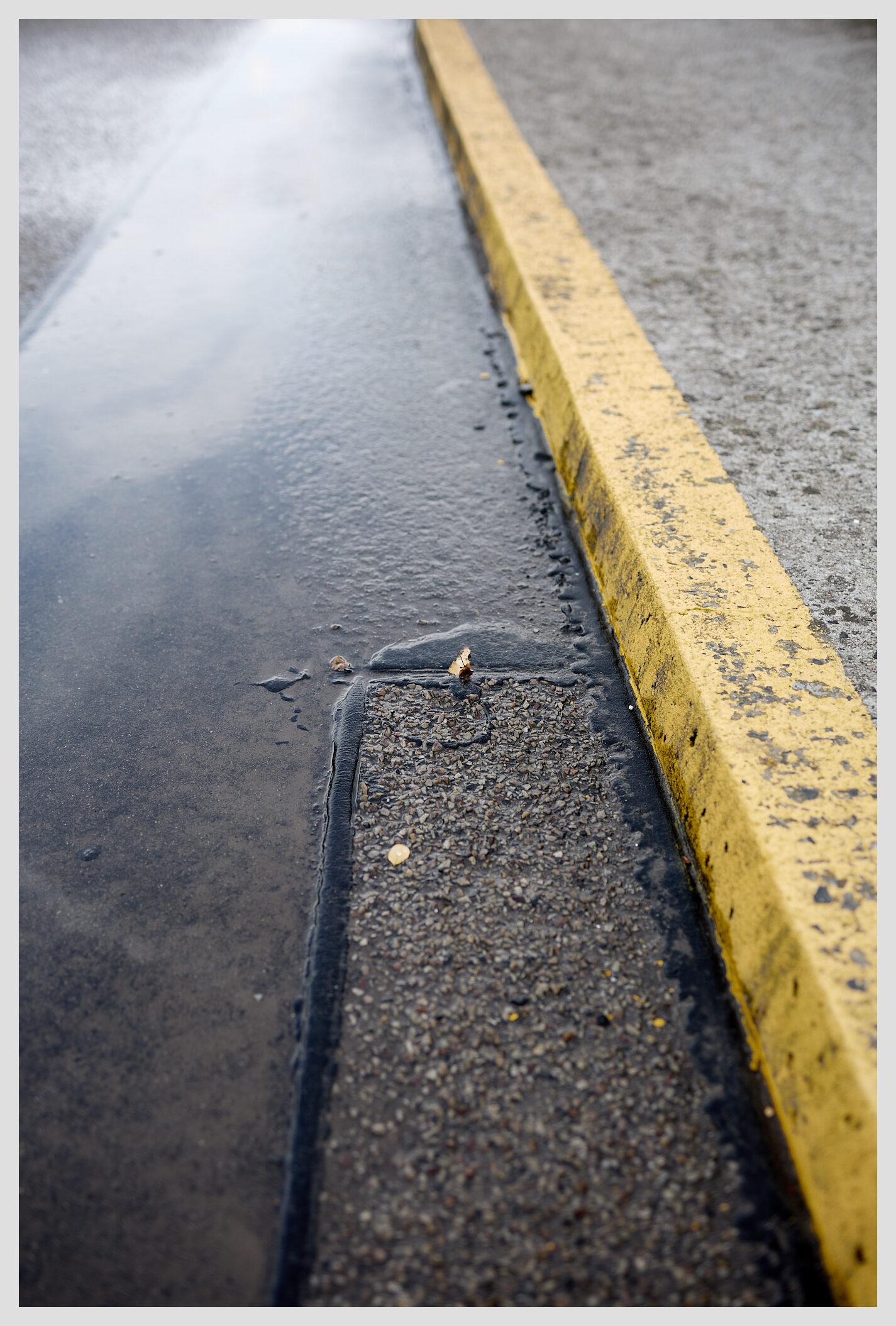 Wide vertical closeup of wet tarmac. A curb painted bright yellow runs along the right side of the frame.