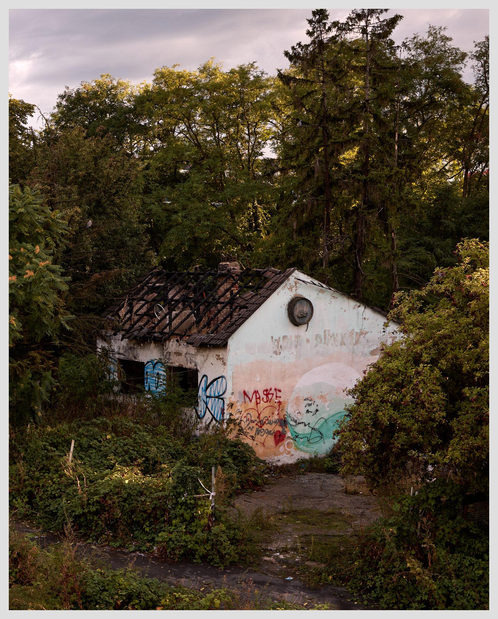 Vertical image of the ruin of a small building whose pointed roof shows heavy fire damage with only charred wooden beams remaining. The outside walls are covered in graffiti. The building is half-hidden between trees.