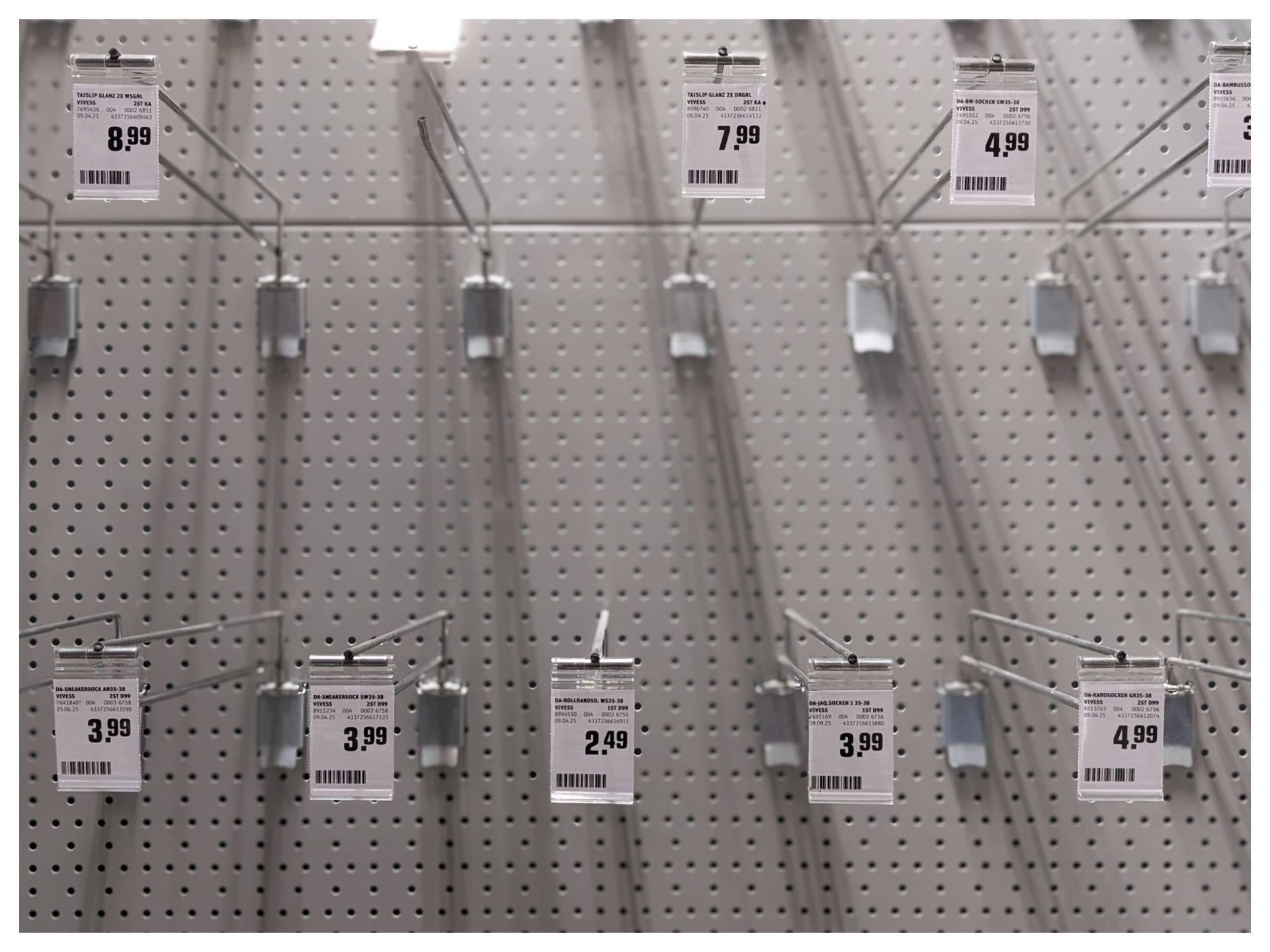 Horizontal black and white image of an empty shelf in a supermarket with price tags attached to metal hangers.