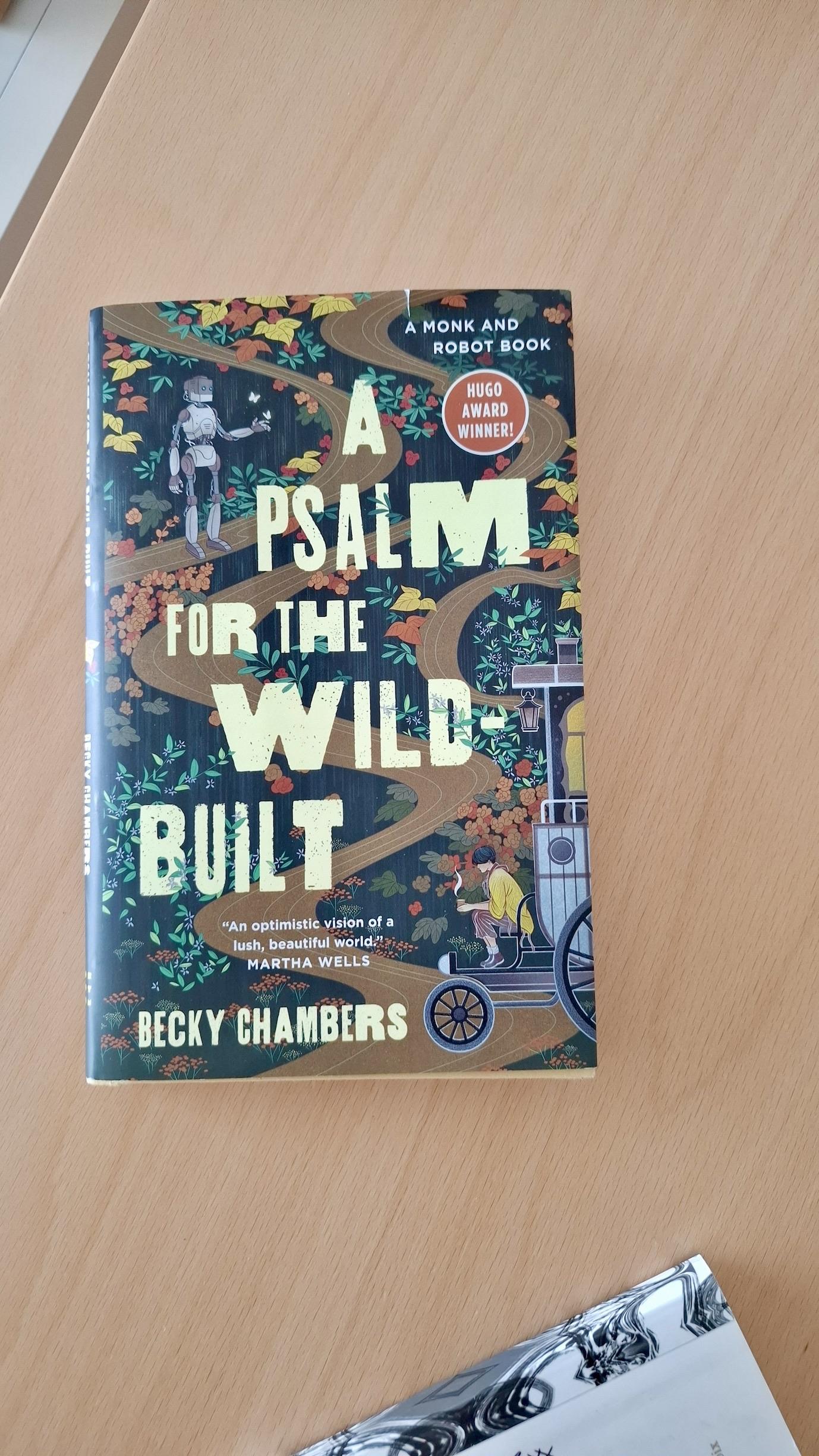 Short version: slim hardback edition of A Psalm for The Wild-Built by Becky Chambers, on a brown table, spine of a notebook nearby. Fuller description: Title and author name in pale yellow block letters, artistic font with letters different sizes and widths. Background: a pretty illustration in autumnal colours of a brown dirt road winding through a stylised forest of yellow and brown and red and green leaves. Bottom right: a figure of a young person with a cup of steaming tea (I know it’s tea because it’s relevant in the book) perced on the edge of a wagon. Above left, a humanoid-shaped robot, gesturing as if inviting you to join it or showing you the way. Other text: A Monk And Robot Book. Hugo Award Winner. Praise quote from Martha Wells: ‘An optimistic vision of a lush, beautiful world.’
