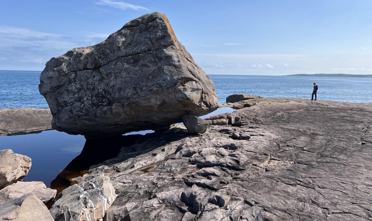 A large erratic (boulder) sits in a still pool of water along a flat rock coast, with a person dwarfed in the background. The sky and ocean are bright blue, with the water blue reflecting a darker blue. The erratic and coast are shades of reddish grey. A spit if land is just visible at the horizon on the right side of the image, just above the silhouettes of the person standing on the coastline. [East Coast Trail Cape Spear Path (NL, Canada) August 2026]