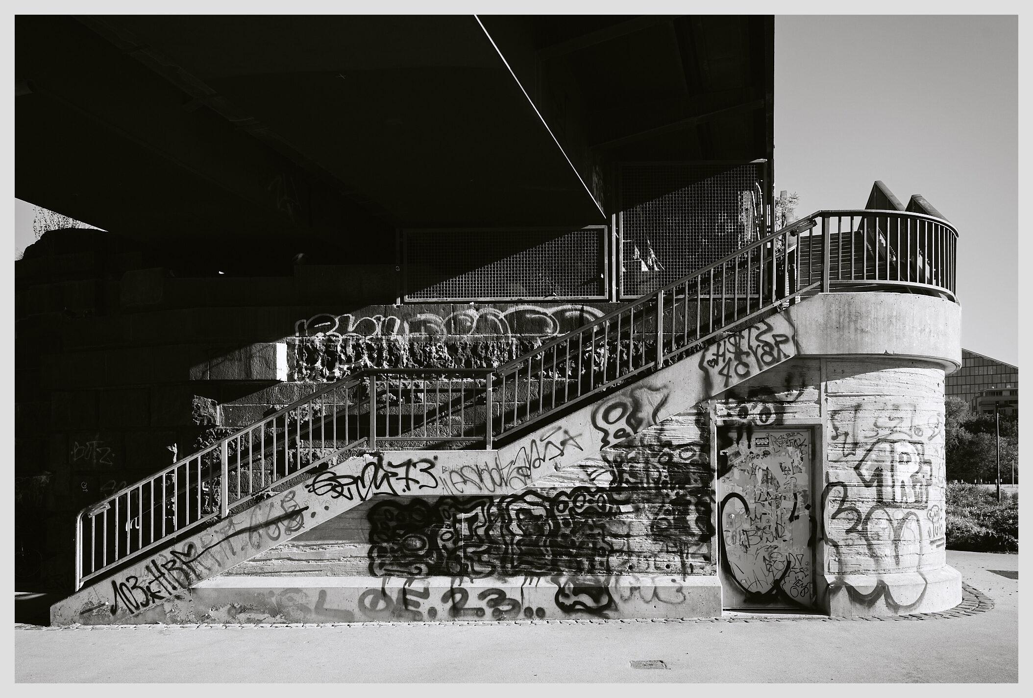 Horizontal black and white image of stairs leading up to a railroad bridge with a footpath. The concrete base of the bridge is covered in graffiti.