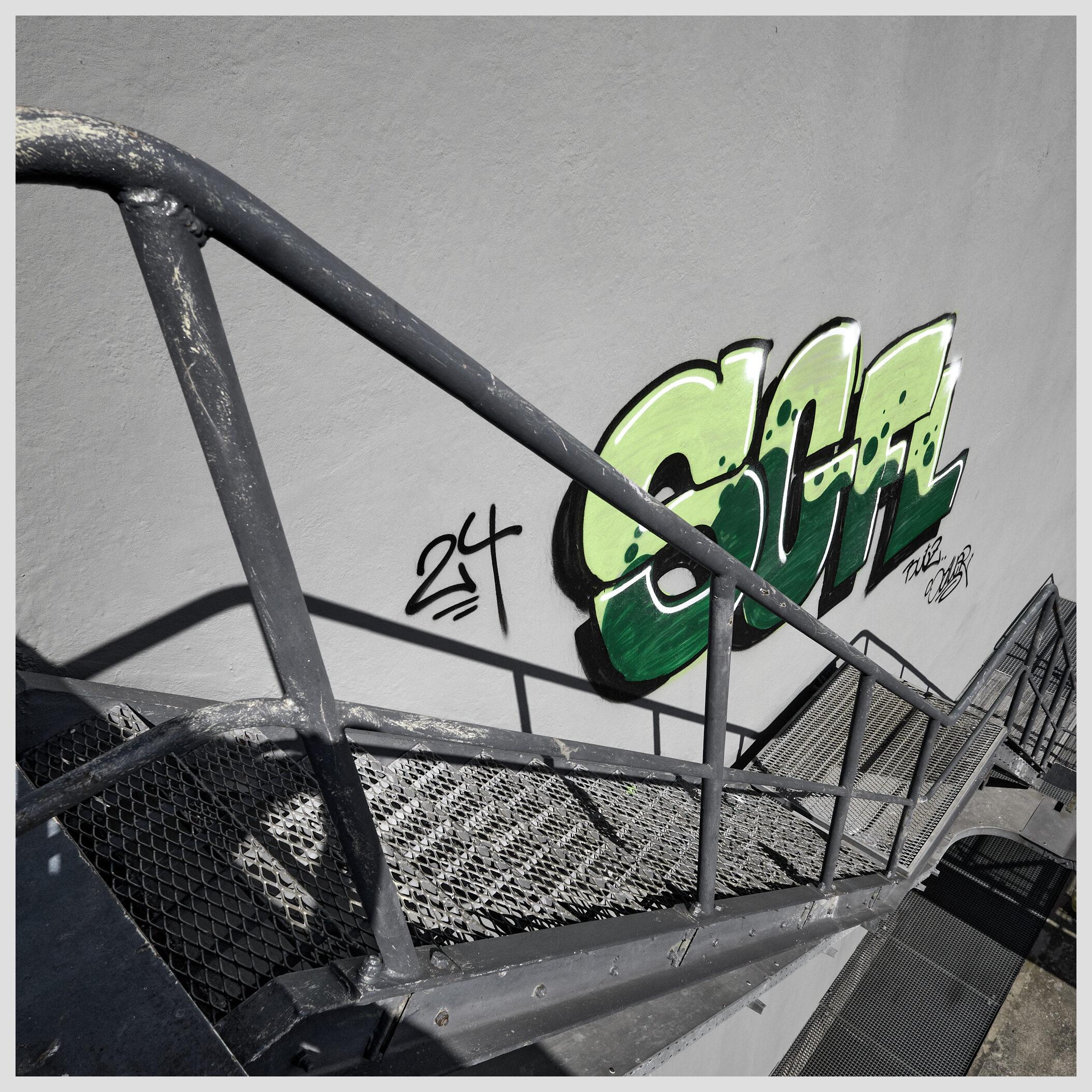 Square image of graffiti on an industrial site. A steep metal staircase is descending next to a gray concrete wall. At the bottom of the stairs, a graffiti in light and dark green tones has been painted on the wall.