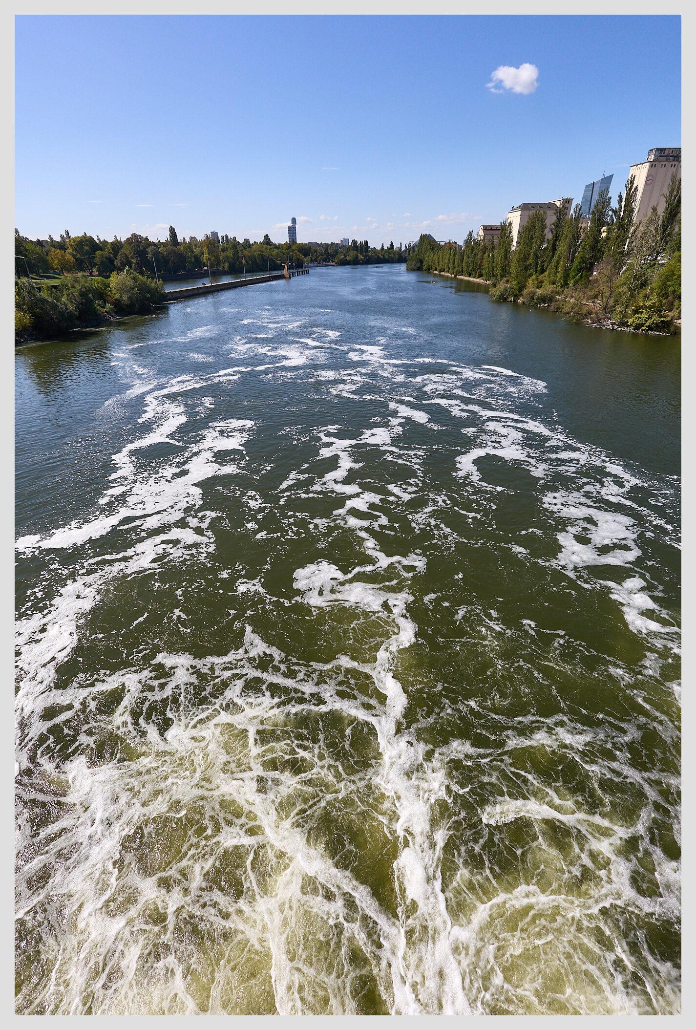 Wide vertical view down a river, taken from the top walk of a floodgate that has recently been opened, causing turbulence in the water below. The riverbank is lined with trees and a few industrial buildings on the right.