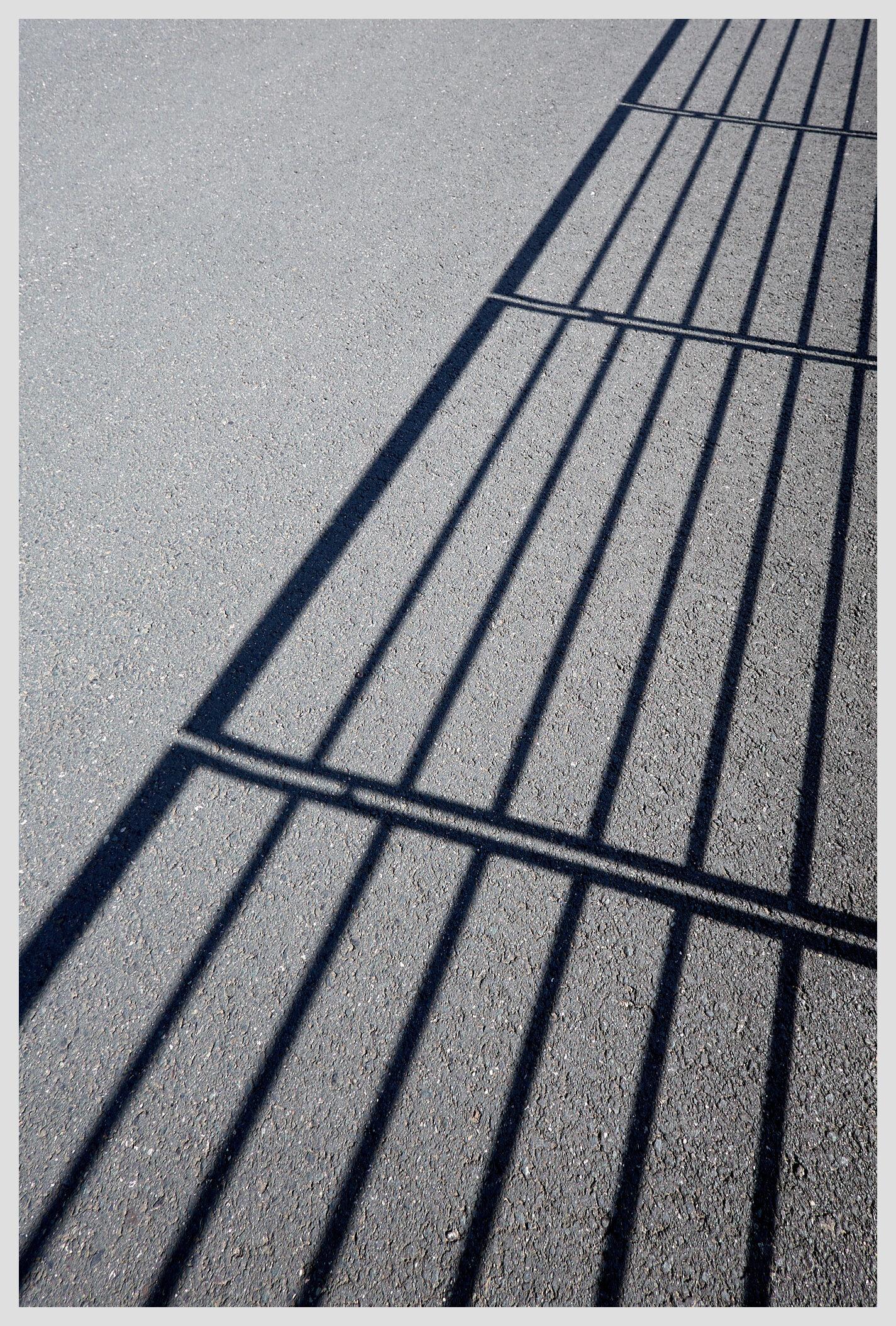 Vertical image of shadow lines cast on a paved footpath by a metal railing.