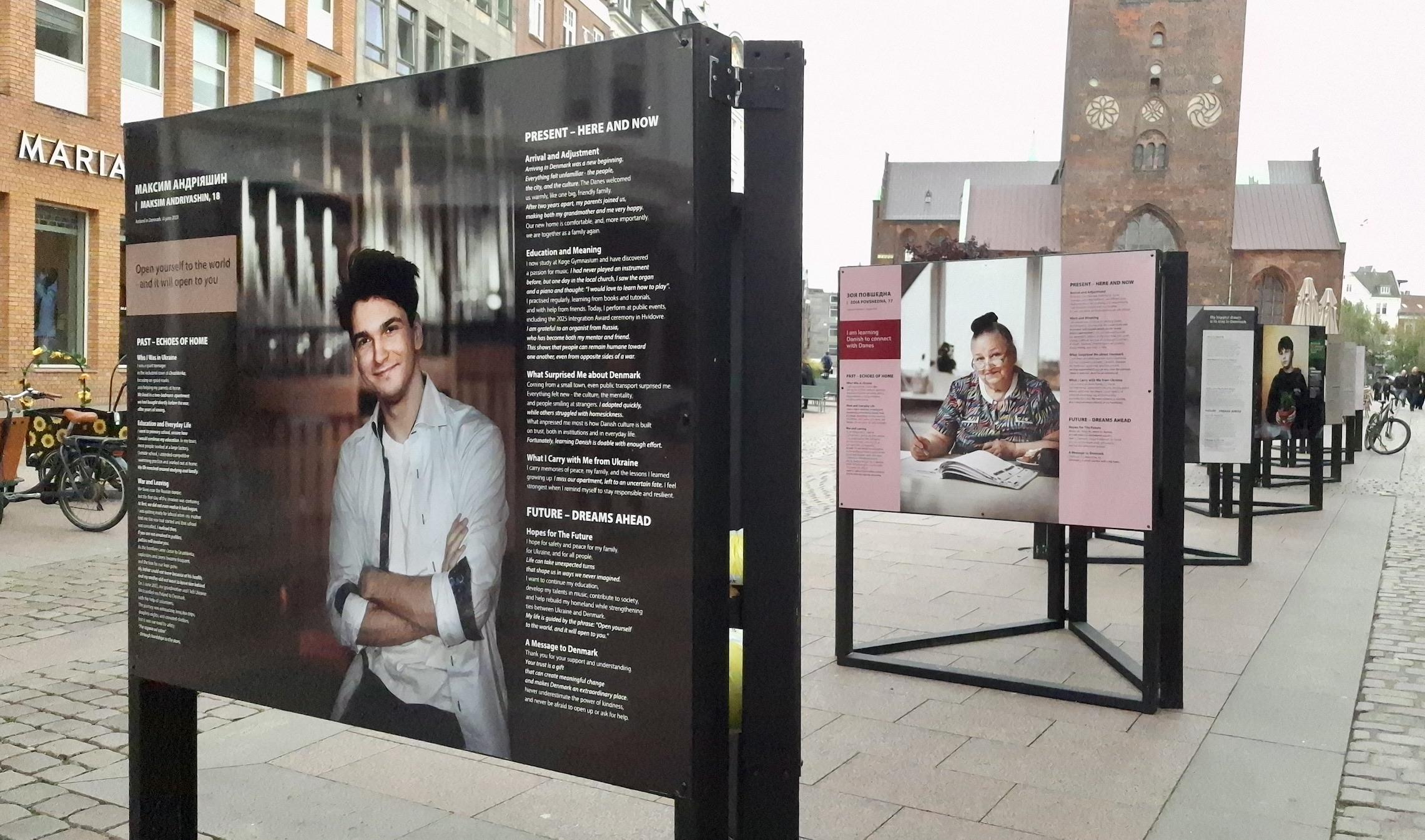 Short version: exhibition billboards. Fuller description: Row of billboards on square, glimpse of cathedral beyond. Street scene. Closest billboard has photo of Maxim, an 18-year-old man with dark hair and white shirt, arms folded, looking at us, smiling. On other billboards, we see an older woman and a young boy. This is from the exhibition in Aarhus, Denmark, on the square by the Domkirken (cathedral). I took the photos in passing in recent weeks. I don’t know if the exhibition is still on show. Short version: exhibition billboards. Fuller description: Row of billboards on square, glimpse of cathedral beyond. Street scene. Closest billboard has photo of Maxim, an 18-year-old man with dark hair and white shirt, arms folded, looking at us, smiling. On other billboards, we see an older woman and a young boy. This is from the exhibition in Aarhus, Denmark, on the square by the Domkirken (cathedral). I took the photos in passing in recent weeks. I don’t know if the exhibition is still on show.