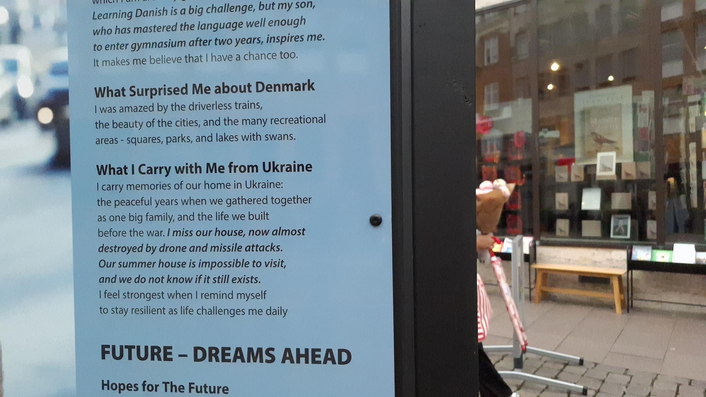 Billboard. Beyond, a person carrying a bouquet of flowers and a bookshop window display. Text on billboard:
‘What surprised me about Denmark’
Driverless trains, beauty of the cities, the many recreational areas - squares, parks, and lakes with swans.
“What I carry with Me From Ukraine”
I carry memories of our home in Ukraine: the peaceful years when we gathered together as one big family, and the life we built before the war. I miss our house, now almost destroyed by drone and missile attacks. our summer house is impossible to visit, and we do not know if it still exists. I feel strongest when I remind myself to stray resilient as life challenges me daily. Billboard. Beyond, a person carrying a bouquet of flowers and a bookshop window display. Text on billboard:
‘What surprised me about Denmark’
Driverless trains, beauty of the cities, the many recreational areas - squares, parks, and lakes with swans.
“What I carry with Me From Ukraine”
I carry memories of our home in Ukraine: the peaceful years when we gathered together as one big family, and the life we built before the war. I miss our house, now almost destroyed by drone and missile attacks. our summer house is impossible to visit, and we do not know if it still exists. I feel strongest when I remind myself to stray resilient as life challenges me daily.