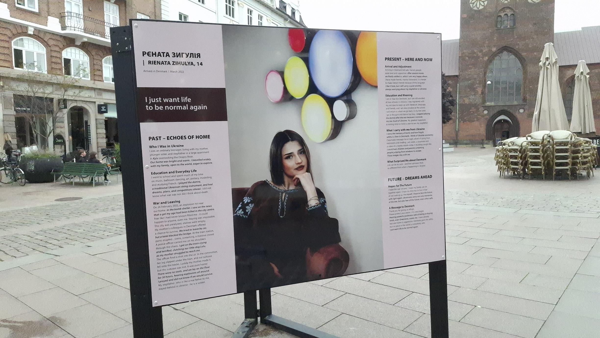 Billboard on city square in Aarhus, Denmark, glimpse of café tables and cathedral beyond. Photo of Rienata, 14 years old, white, seated, hand on chin, gazing at us, coloured wall decorations behind her. She is in dark clothes and has straight dark hair. Text around it, sorry, just an extract here due to length:
‘Who I was in Ukraine’ - an ordinary teenager in Kyiv. Travelled widely with family, open to world, eager to explore.
Everyday life: school, ballroom dancing, art, pottery, modelling, learning French.
War and Leaving: On 24 February 2022, an explosion hit near our home. Girl my age killed in city centre. Fear like I have never known filled me. Staying was impossible. At train station, panic erupted - sirens, screaming, a massive crowd. A police officer carried me on his shoulders through the chaos. I got on the train crying and terrified, clutching our little dog Lulu, as my mother struggled to reached me. The officer fired a shot into the air. In the commotion, her leg slipped under the train, and our suitcase fell onto the tracks. Luckily my mother made it, but hte suitcase was lost. It was pure horror. There were no seats, and we lay on the floor for 30 hours, hearing explosions all around. I prayed and did now know if we would survive. My stepfather, who is like a real father to me, stayed behind in Ukraine - he is a soldier. Billboard on city square in Aarhus, Denmark, glimpse of café tables and cathedral beyond. Photo of Rienata, 14 years old, white, seated, hand on chin, gazing at us, coloured wall decorations behind her. She is in dark clothes and has straight dark hair. Text around it, sorry, just an extract here due to length:
‘Who I was in Ukraine’ - an ordinary teenager in Kyiv. Travelled widely with family, open to world, eager to explore.
Everyday life: school, ballroom dancing, art, pottery, modelling, learning French.
War and Leaving: On 24 February 2022, an explosion hit near our home. Girl my age killed in city centre. Fear like I have never known filled me. Staying was impossible. At train station, panic erupted - sirens, screaming, a massive crowd. A police officer carried me on his shoulders through the chaos. I got on the train crying and terrified, clutching our little dog Lulu, as my mother struggled to reached me. The officer fired a shot into the air. In the commotion, her leg slipped under the train, and our suitcase fell onto the tracks. Luckily my mother made it, but hte suitcase was lost. It was pure horror. There were no seats, and we lay on the floor for 30 hours, hearing explosions all around. I prayed and did now know if we would survive. My stepfather, who is like a real father to me, stayed behind in Ukraine - he is a soldier.